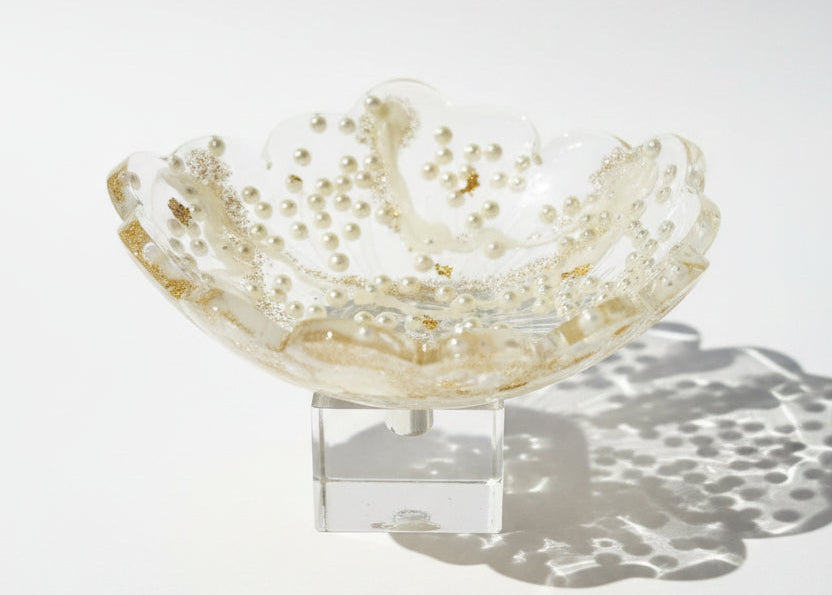 A scalloped clear resin dish filled with small white pearls and gold flakes, photographed on a white surface with strong sunlight and shadows.