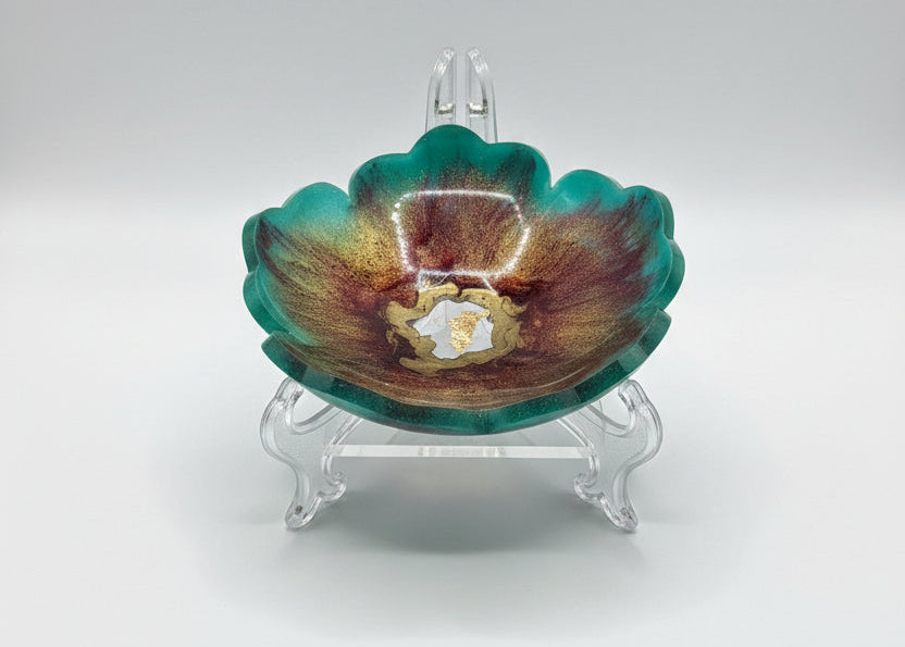 A small flower-shaped resin dish with a marbled pattern of green, brown, and gold, containing a small central gold inlay, photographed on a white sheet of paper.