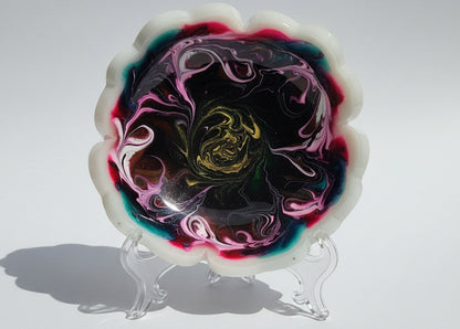 A flower-shaped resin tray or mold with a scalloped white edge, filled with a marbled swirl pattern of black, pink, purple, teal, and yellow, photographed on a white sheet of paper.