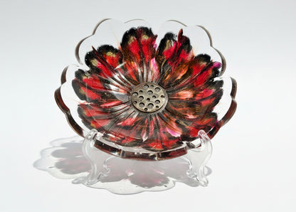 A flower-shaped clear glass bowl with a red, pink, and black painted floral design, set against a white background with strong natural sunlight and shadows.
