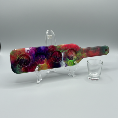 Colorful shot glass paddle for four shot glasses on a stand next to a clear shot glass on a gray background.